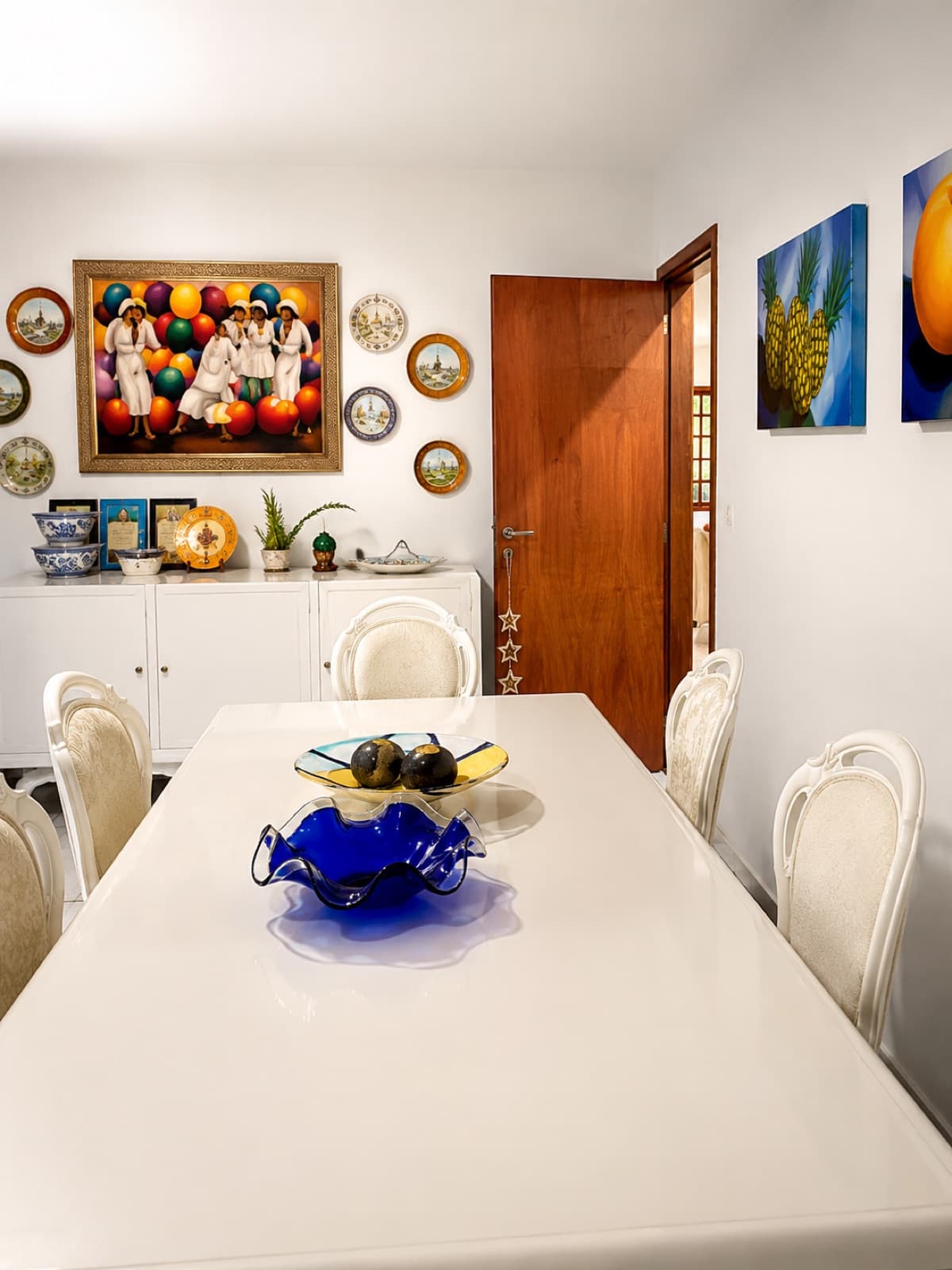 Sala de jantar clara com mesa branca e quadros coloridos nas paredes