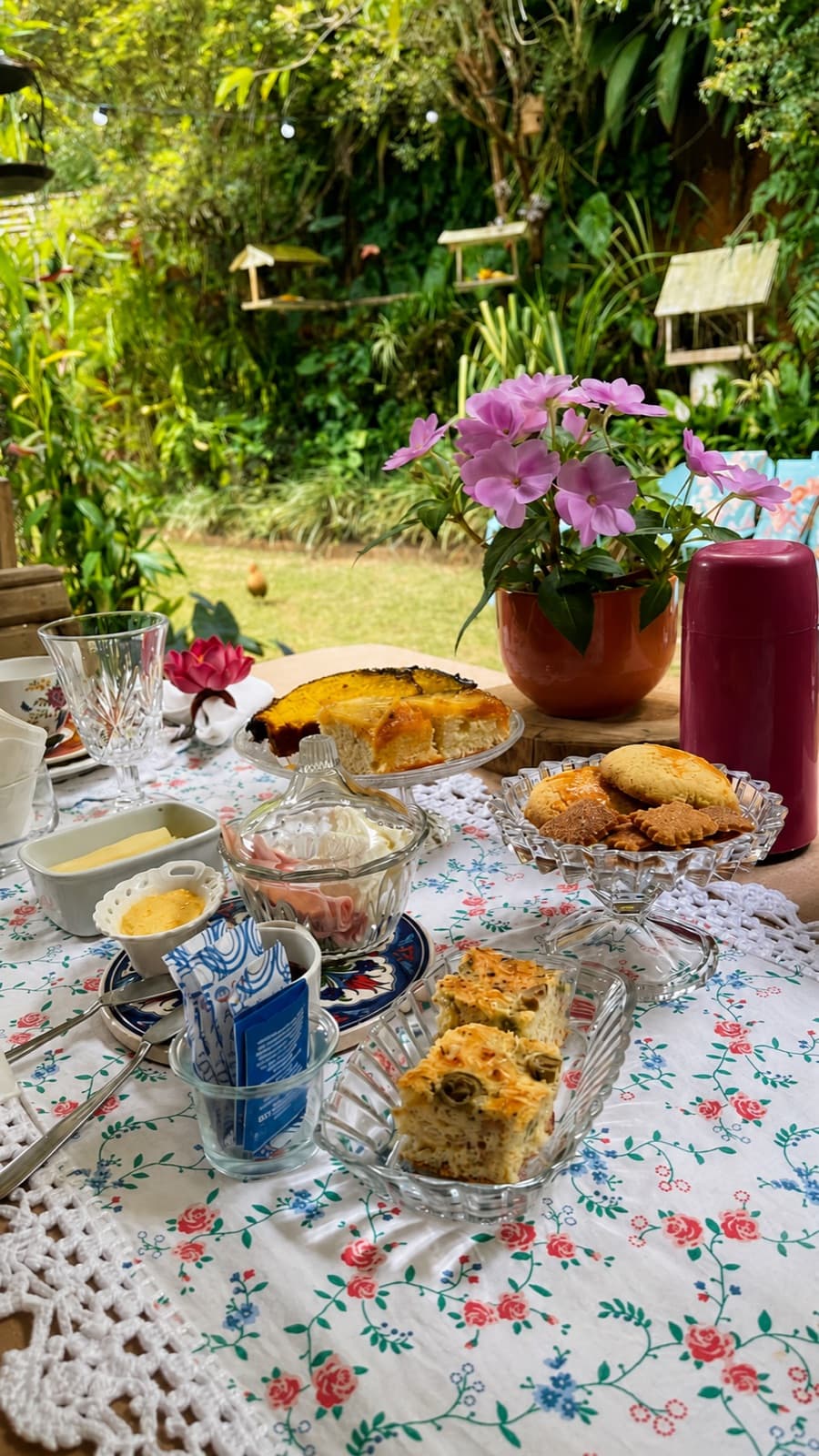 Mesa posta com toalha floral, bolos caseiros e vista para o jardim